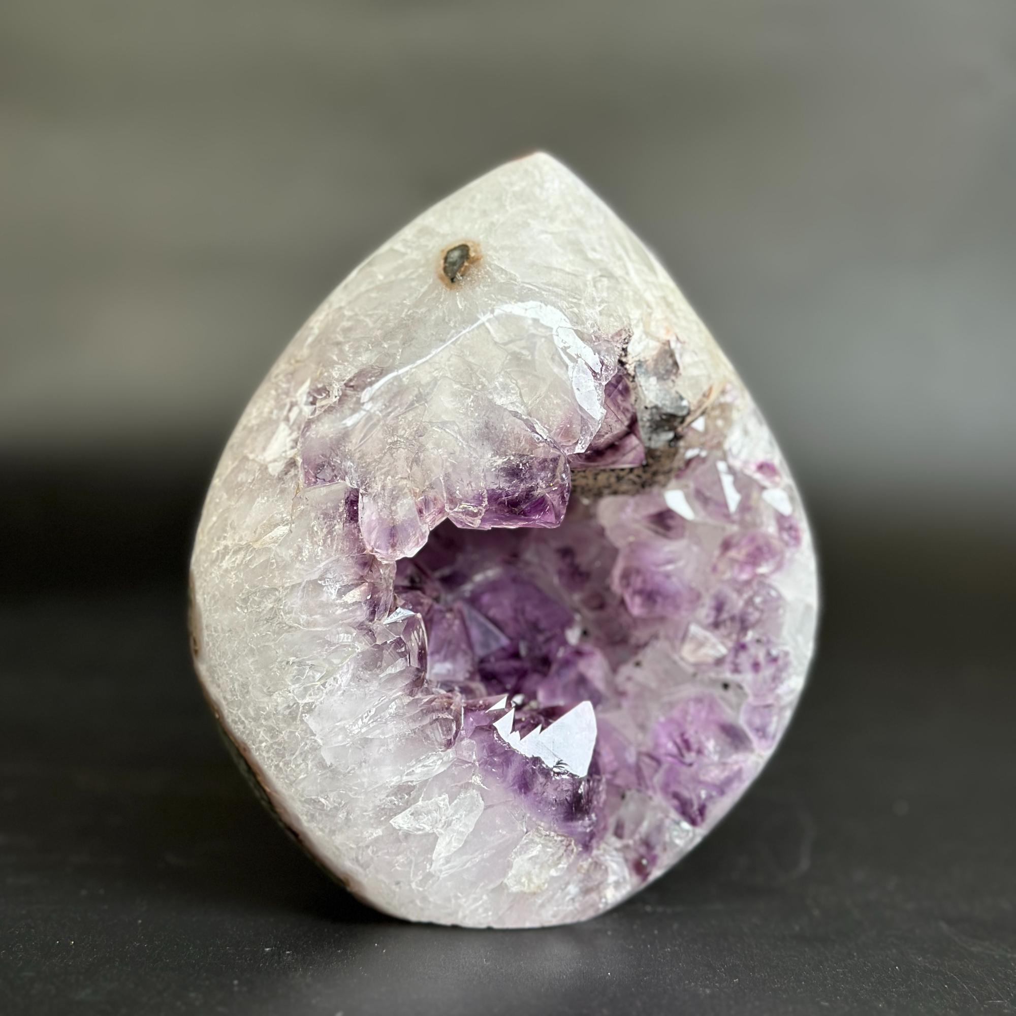 A polished natural amethyst geode specimen with a white and purple crystal interior, photographed on a dark gray surface against a blurred gray background.
