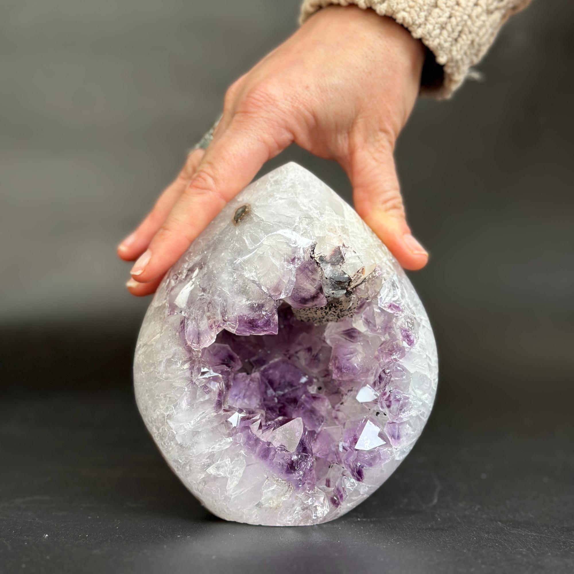 A hand holding a large teardrop-shaped amethyst geode crystal with a white outer layer and a purple crystal cavity, set against a dark gray background.
