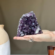 Deep purple Uruguayan amethyst geode crystal cluster held in a hand, next to a beige vase.