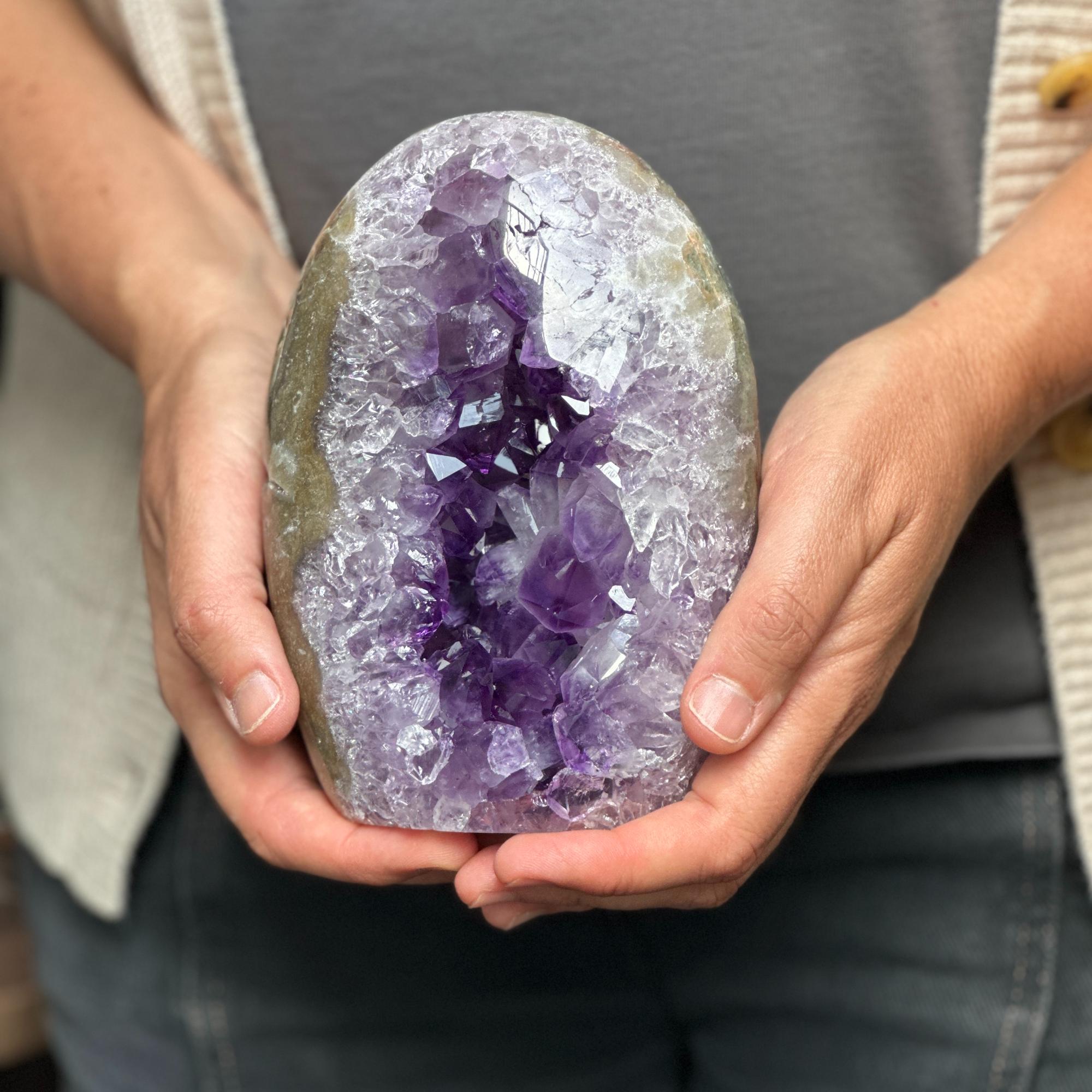 Hands hold a vibrant deep purple amethyst geode, egg-shaped with sparkling crystals and natural agate edges.