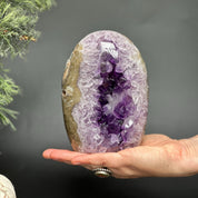 Hand holding a 6-inch tall, egg-shaped Uruguayan amethyst geode with deep purple crystals and agate edges.