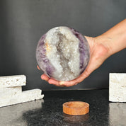 Hand holds a 4-inch amethyst geode sphere with sparkling white quartz crystals, next to its carob wood stand.
