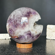 4-inch polished purple amethyst quartz crystal sphere with a sparkling white geode opening, on a round carob wood base.