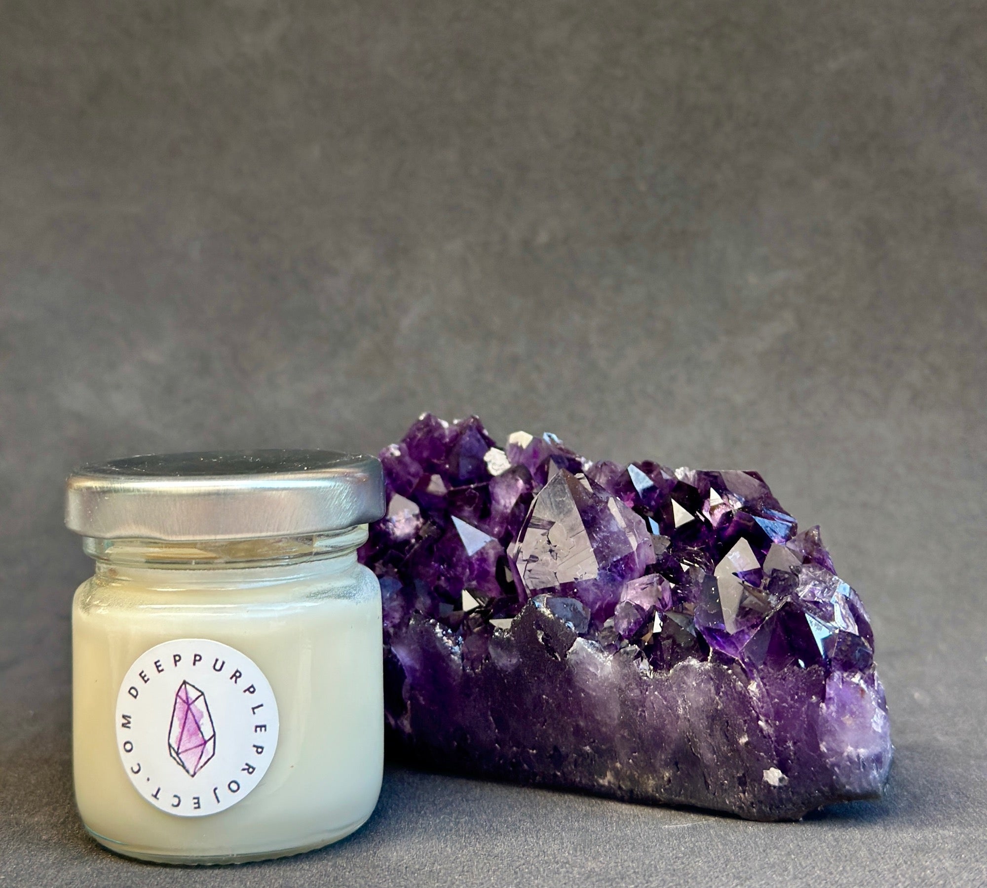 Small candle in a glass jar with a purple crystal formation on a gray background