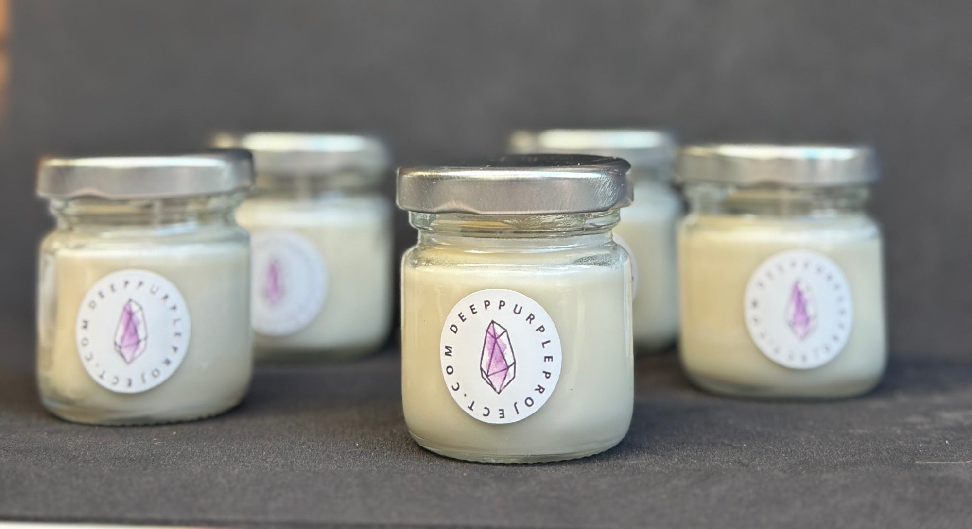 Five small jars with candles inside, each labeled with a circular sticker on a dark background.