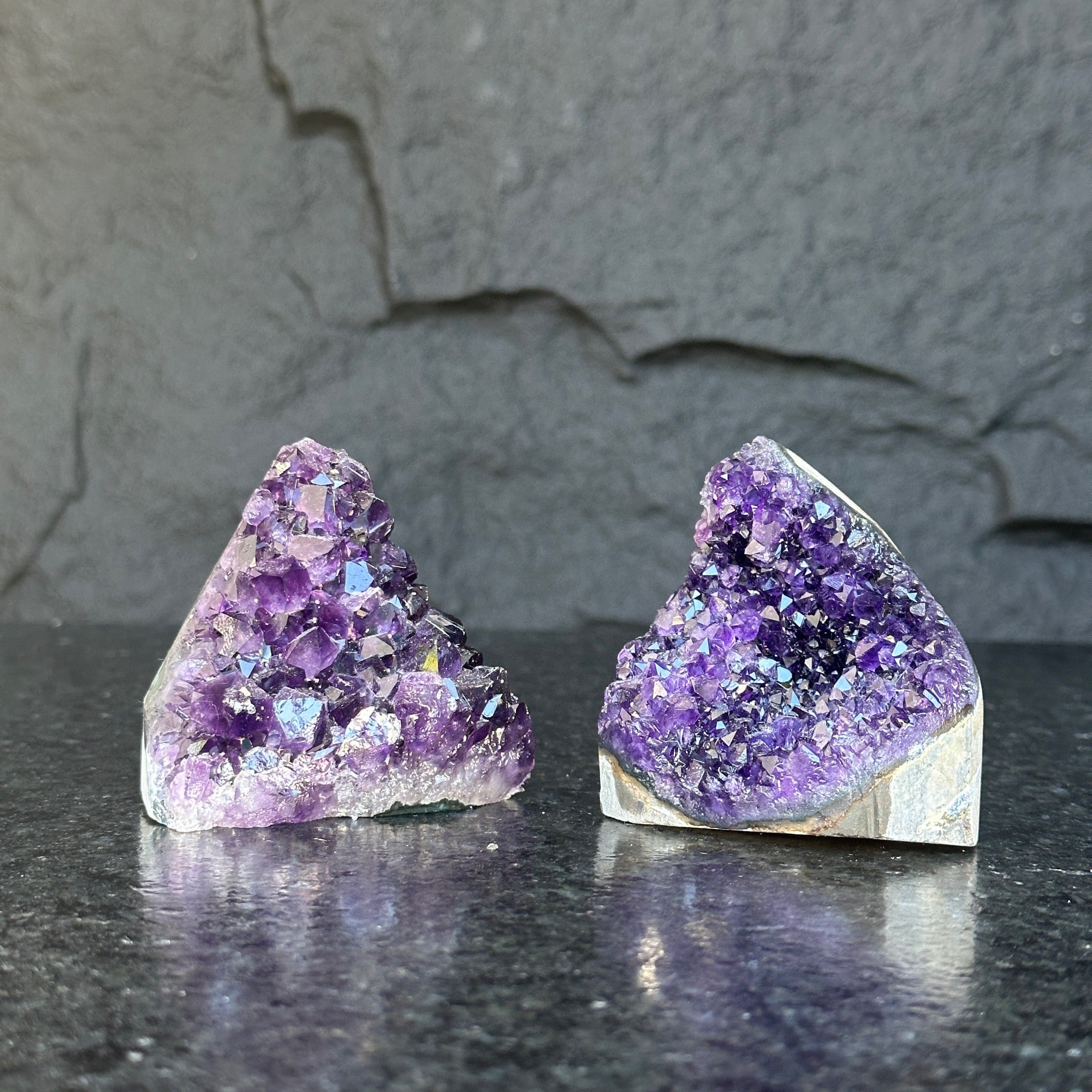 Two small, raw Uruguayan amethyst geode clusters with deep purple crystals on a dark surface.
