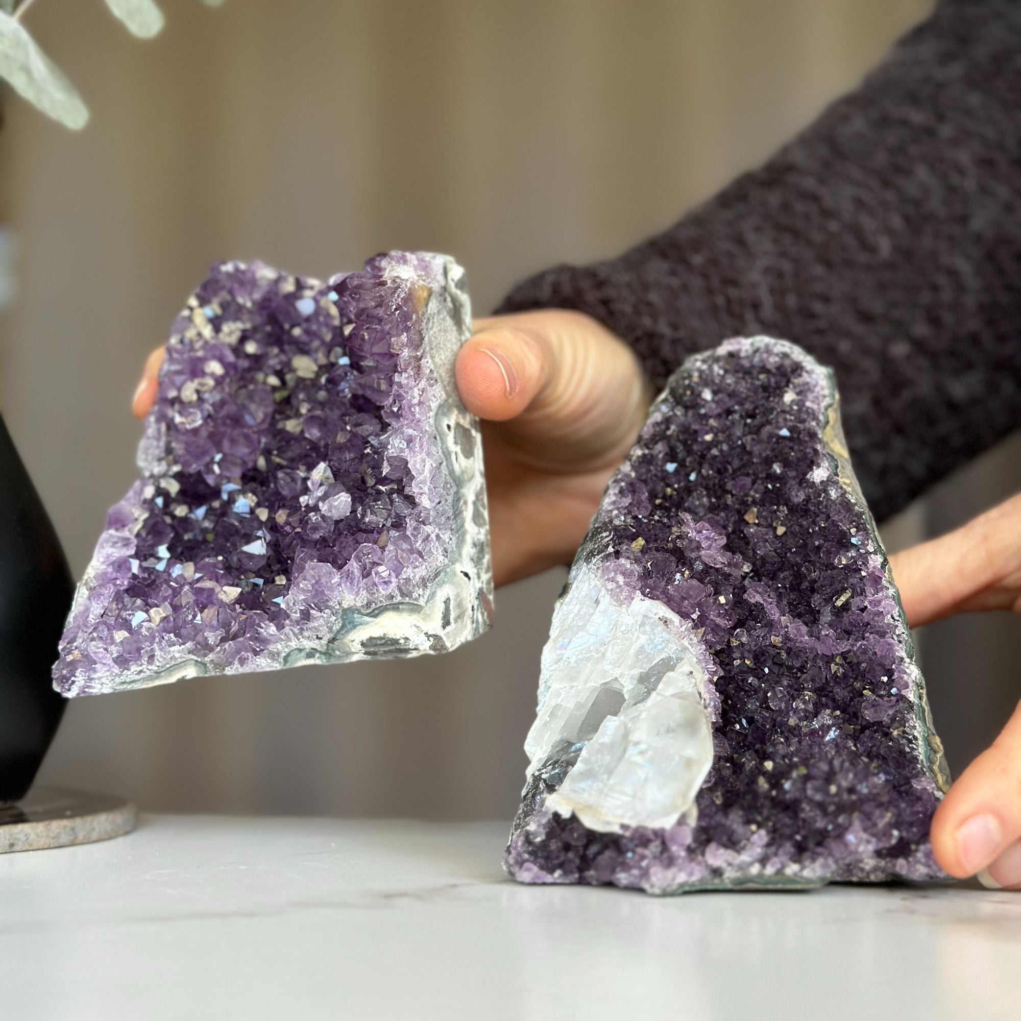 Two deep purple amethyst geodes with calcite formations, held by hands.