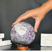 Polished purple amethyst gem stone sphere with white inclusions on a carob wood base, held by a hand.