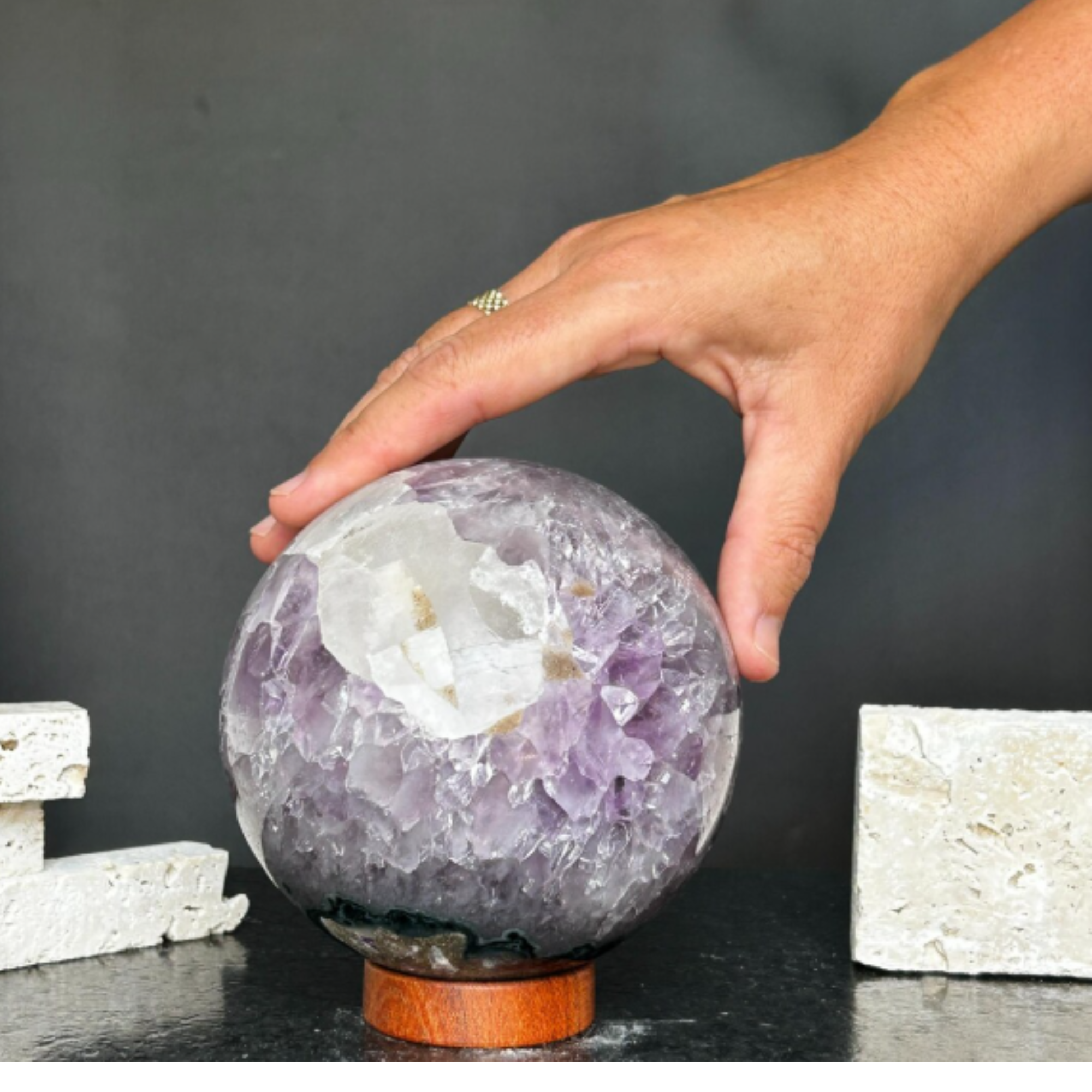 Polished purple amethyst gem stone sphere with white inclusions on a carob wood base, held by a hand.
