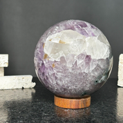 Large purple amethyst gemstone sphere with clear crystal formations on a round carob wood base.