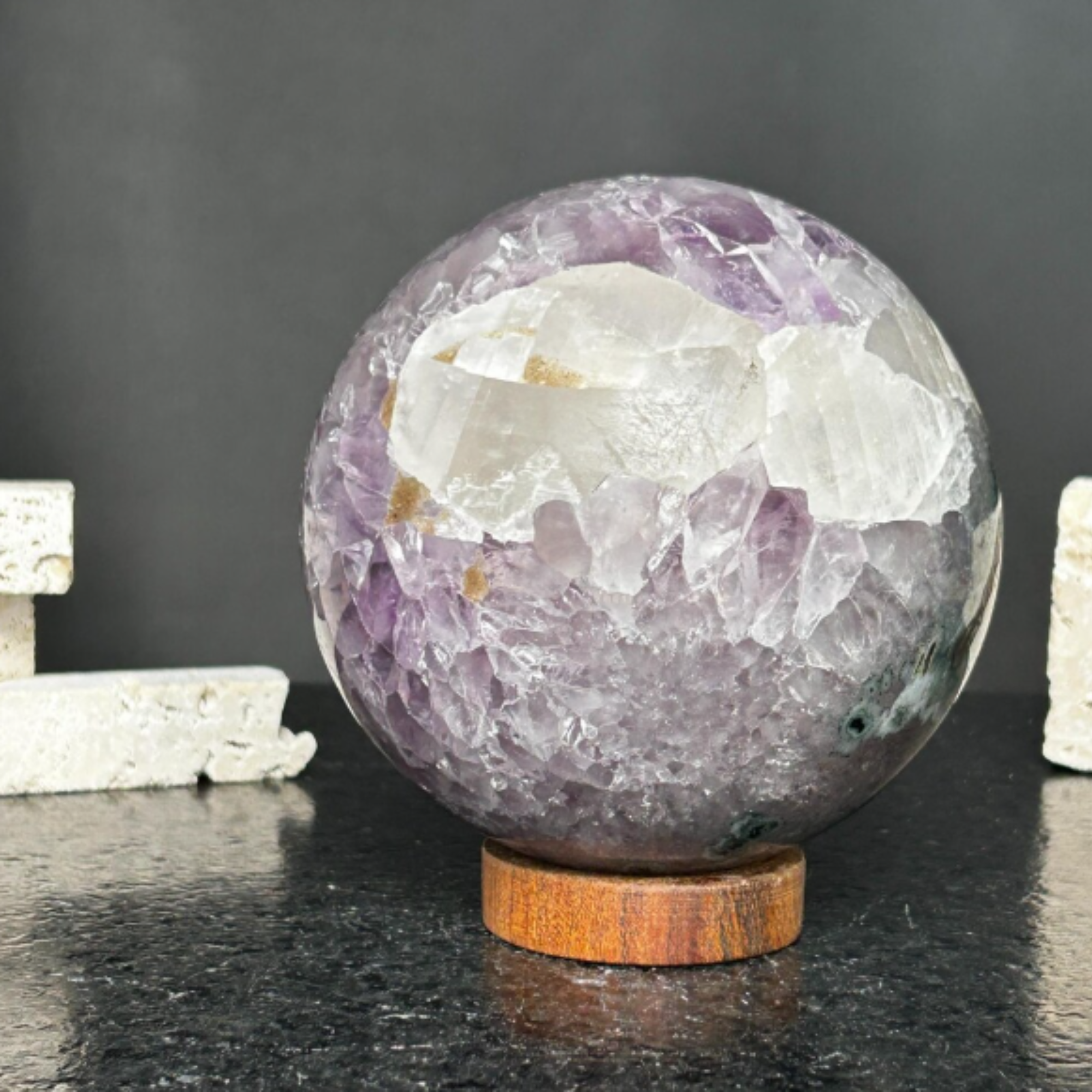 Large purple amethyst gemstone sphere with clear crystal formations on a round carob wood base.