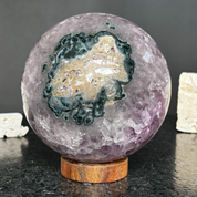 Polished 4-inch purple amethyst sphere with a striking green and gold crystal geode formation, on a carob wood base.