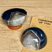 Two polished agate half-spheres on wooden stands, placed on a wooden surface with a 'Thinking of You' greeting card in the background.