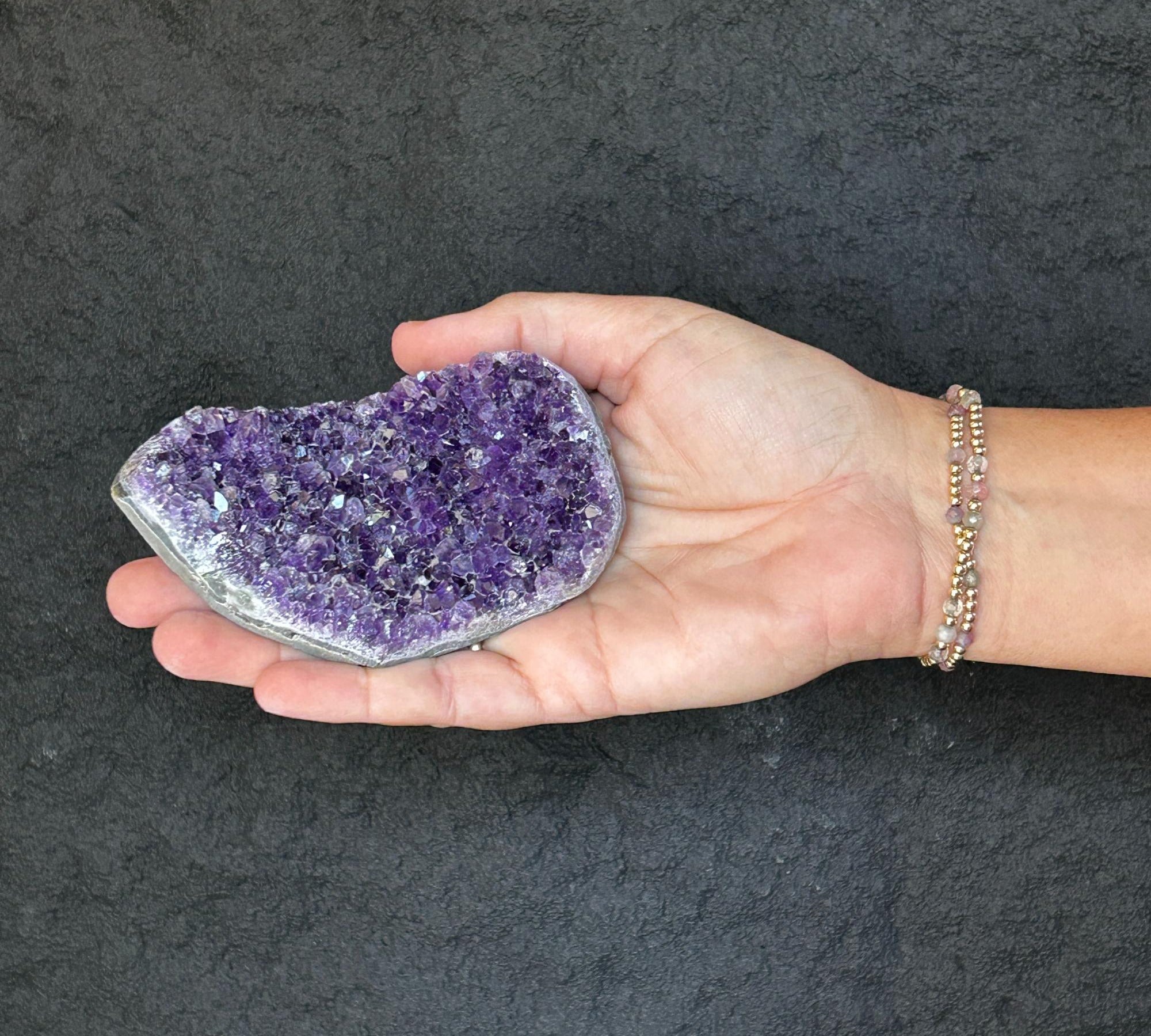 Hand holding a flat, raw purple amethyst crystal cluster geode, 2.5x4 inches, with a beaded bracelet.