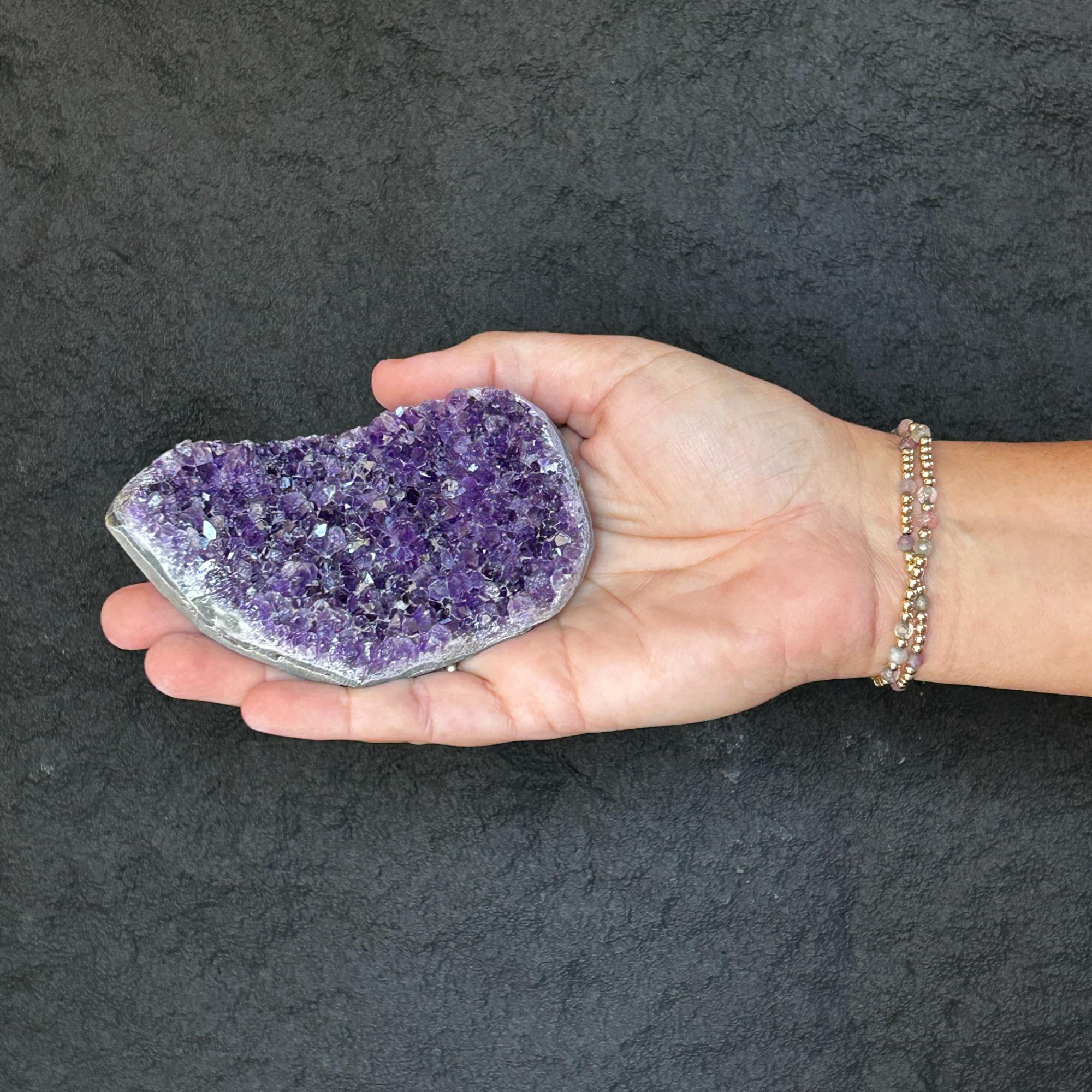 Hand holding a flat, raw purple amethyst crystal cluster geode, 2.5x4 inches, with a beaded bracelet.