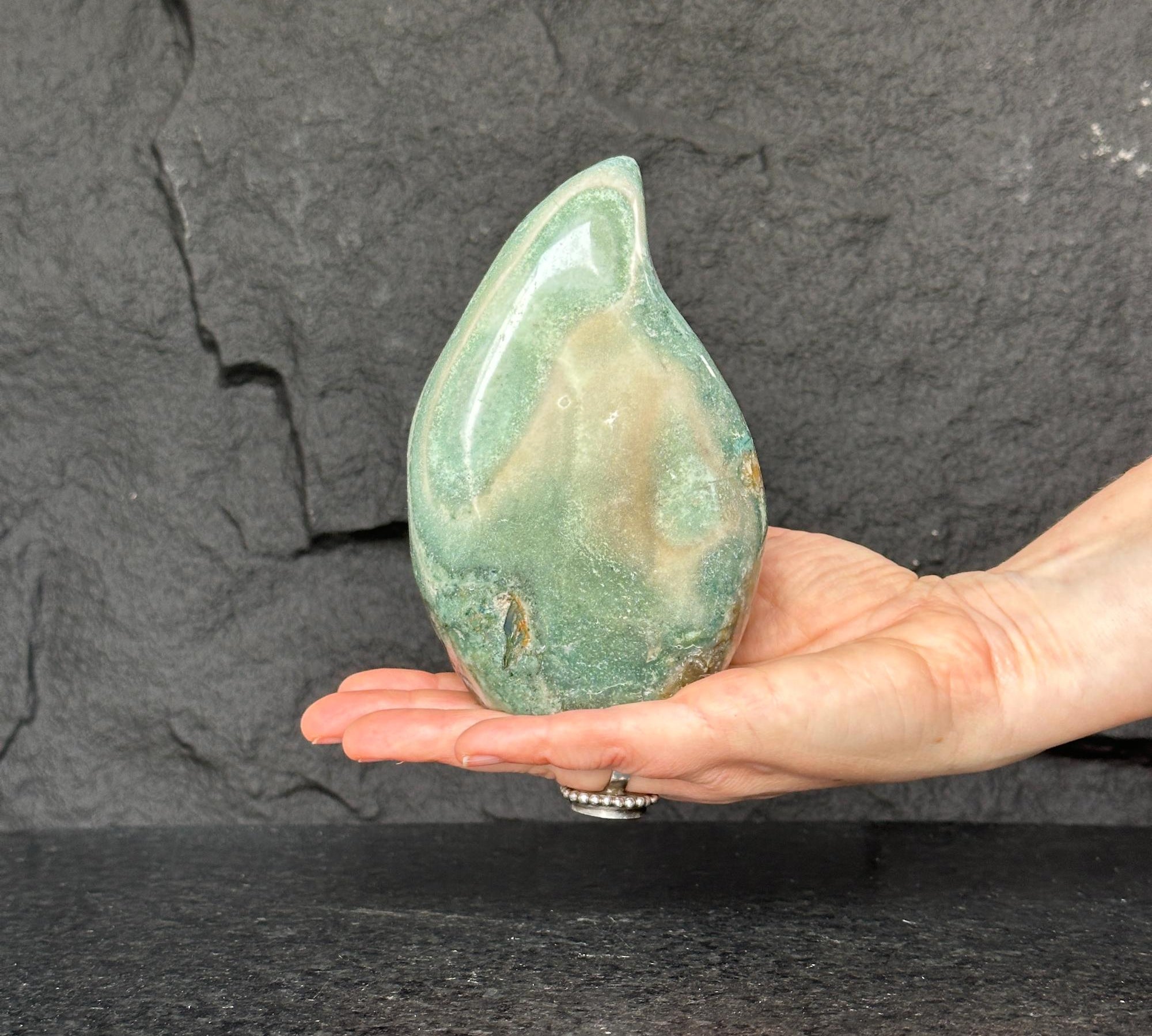 Hand holding a large, polished green jasper flame gemstone with natural brown and cream patterns.