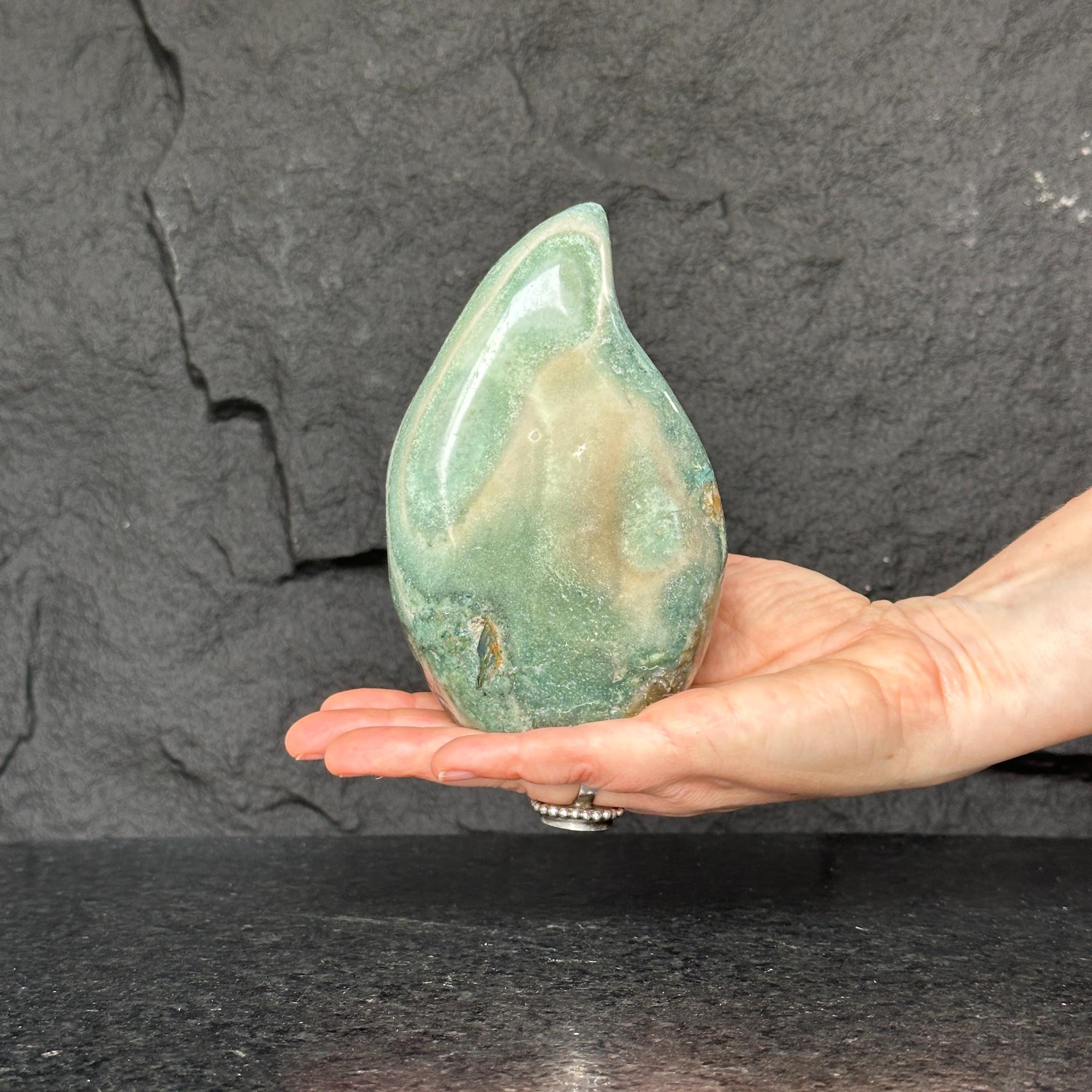 Hand holding a large, polished green jasper flame gemstone with natural brown and cream patterns.