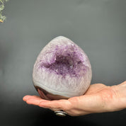 Hand holding a polished, egg-shaped deep purple amethyst geode with white and brown agate edges.