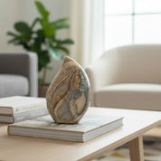A decorative teardrop-shaped jasper stone sculpture with natural marbled patterns in beige, brown, and gray, displayed on a stack of books on a light wood coffee table in a modern living room setting.