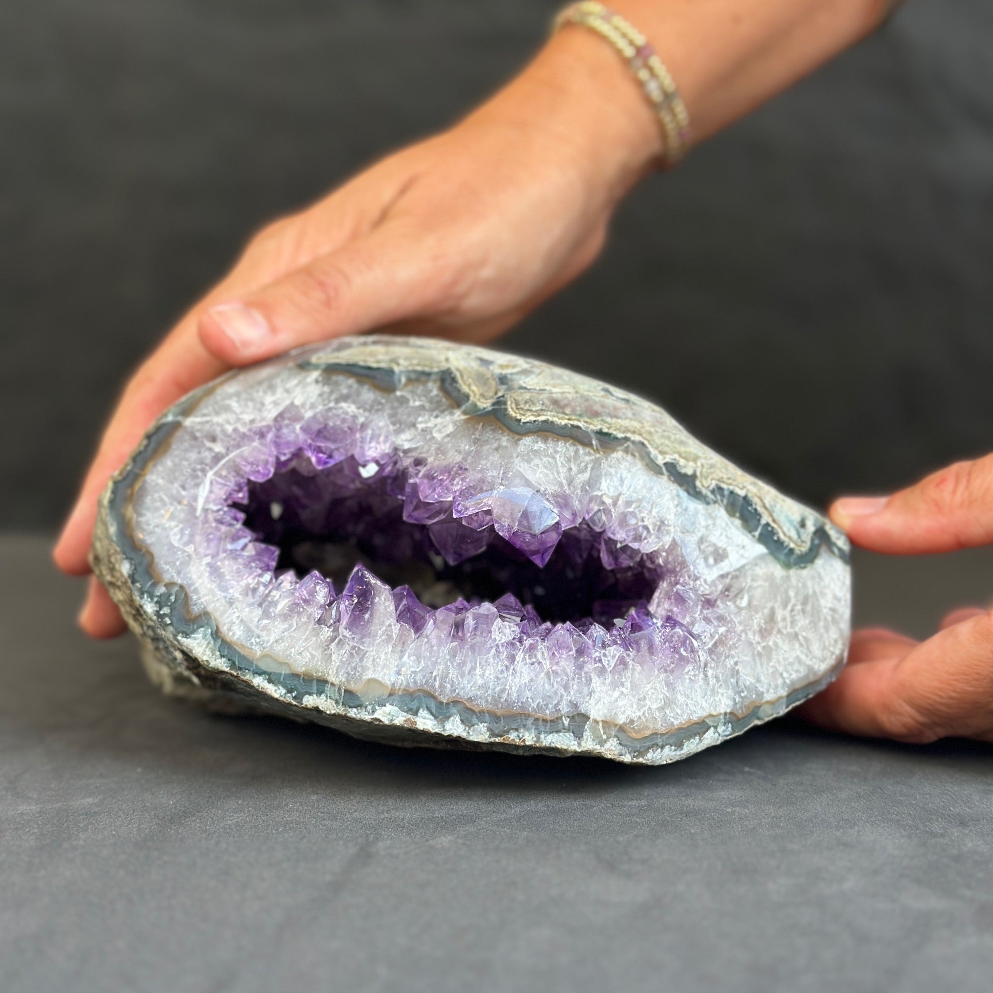 Person holding a large amethyst geode with visible purple interior on a gray surface.