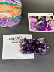 Amethyst crystal on a card with motivational text, next to a colorful amethyst crystal and a card with a purple crystal image on a gray surface.