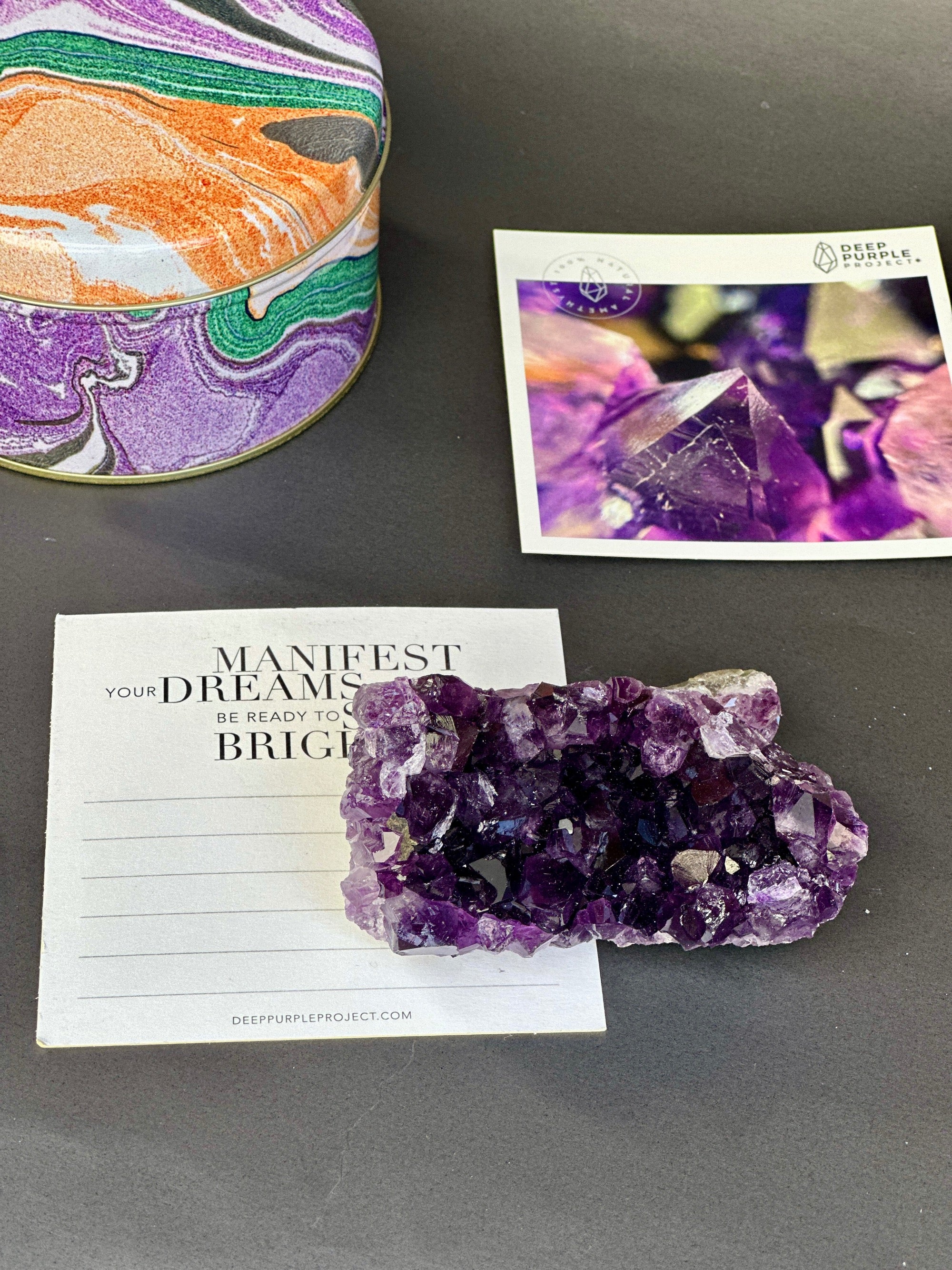 Amethyst crystal on a card with motivational text, next to a colorful amethyst crystal and a card with a purple crystal image on a gray surface.