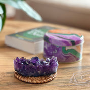 Amethyst crystal on a woven coaster with a candle and book in the background on a wooden surface.