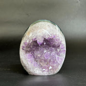 A polished amethyst geode specimen with purple crystal clusters inside a white and gray outer matrix, photographed on a dark gray surface against a dark gray background.