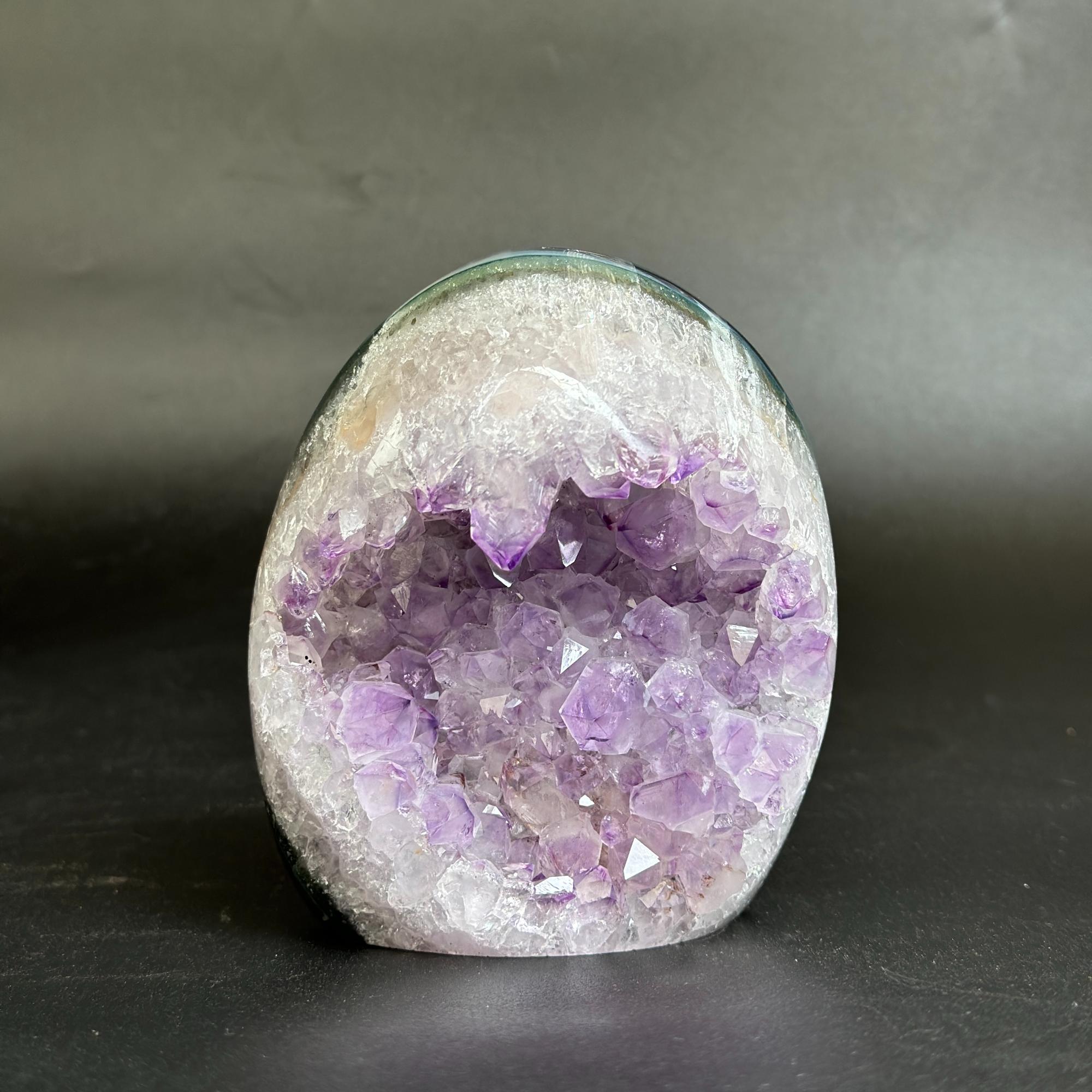 A polished amethyst geode specimen with purple crystal clusters inside a white and gray outer matrix, photographed on a dark gray surface against a dark gray background.