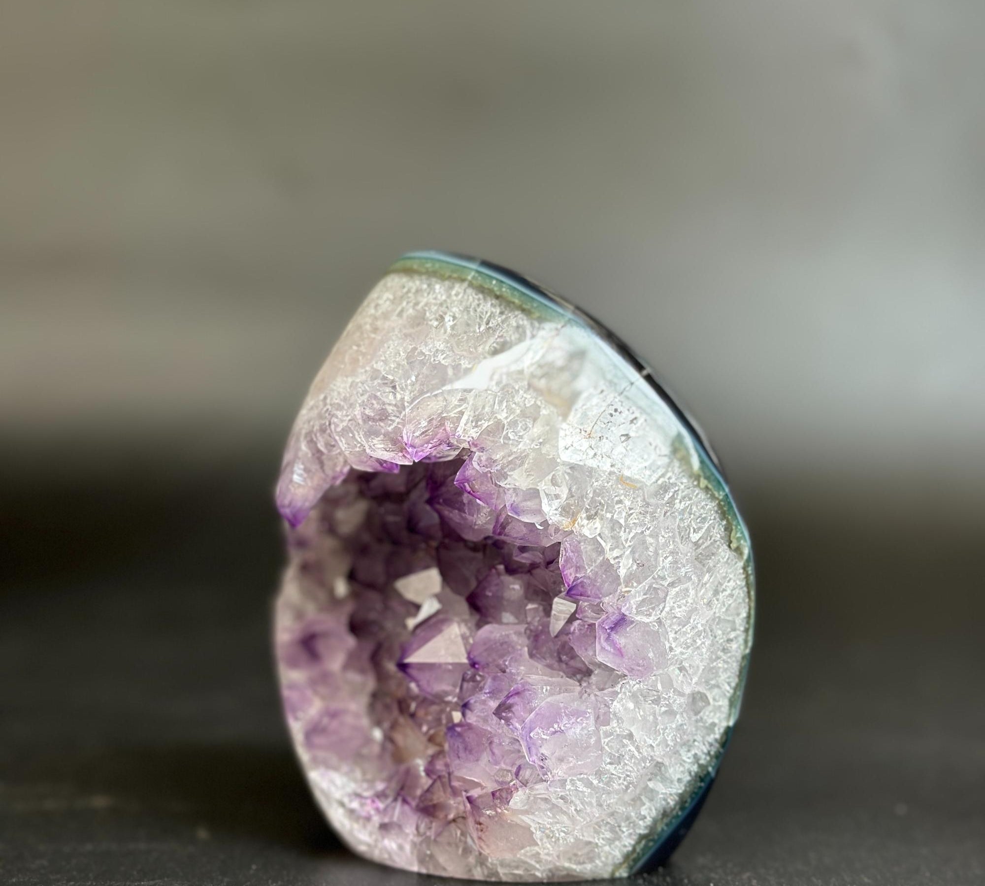 A natural amethyst geode specimen with a white crystalline outer layer and a purple crystal interior, photographed on a dark surface against a blurred gray background.