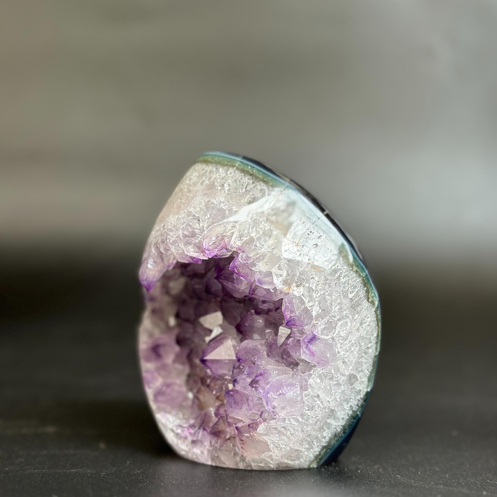 A natural amethyst geode specimen with a white crystalline outer layer and a purple crystal interior, photographed on a dark surface against a blurred gray background.