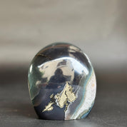 A polished oval agate crystal stone with a dark navy and black body, light green bands, and metallic gold inclusions, standing on a dark surface against a blurred gray background.