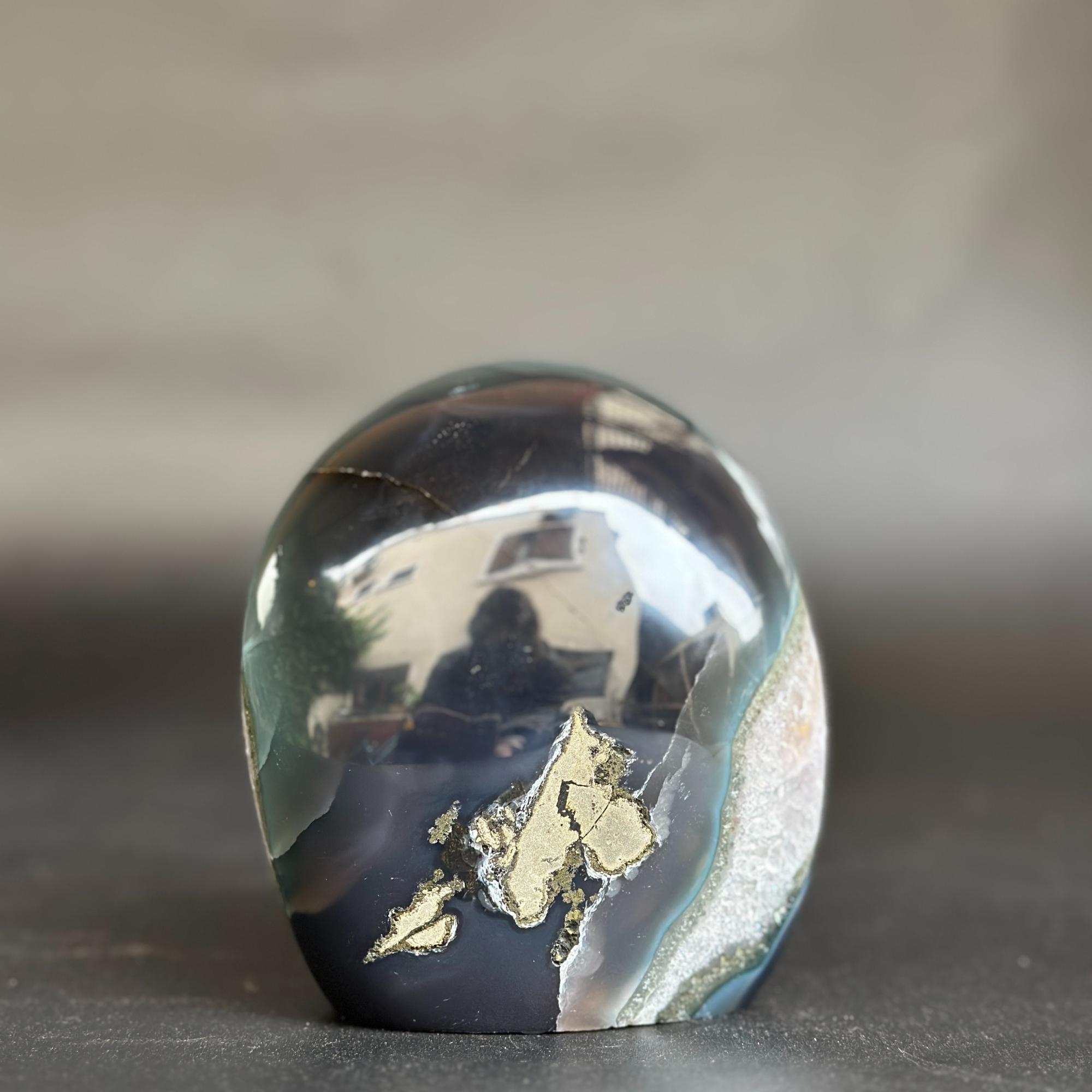 A polished oval agate crystal stone with a dark navy and black body, light green bands, and metallic gold inclusions, standing on a dark surface against a blurred gray background.
