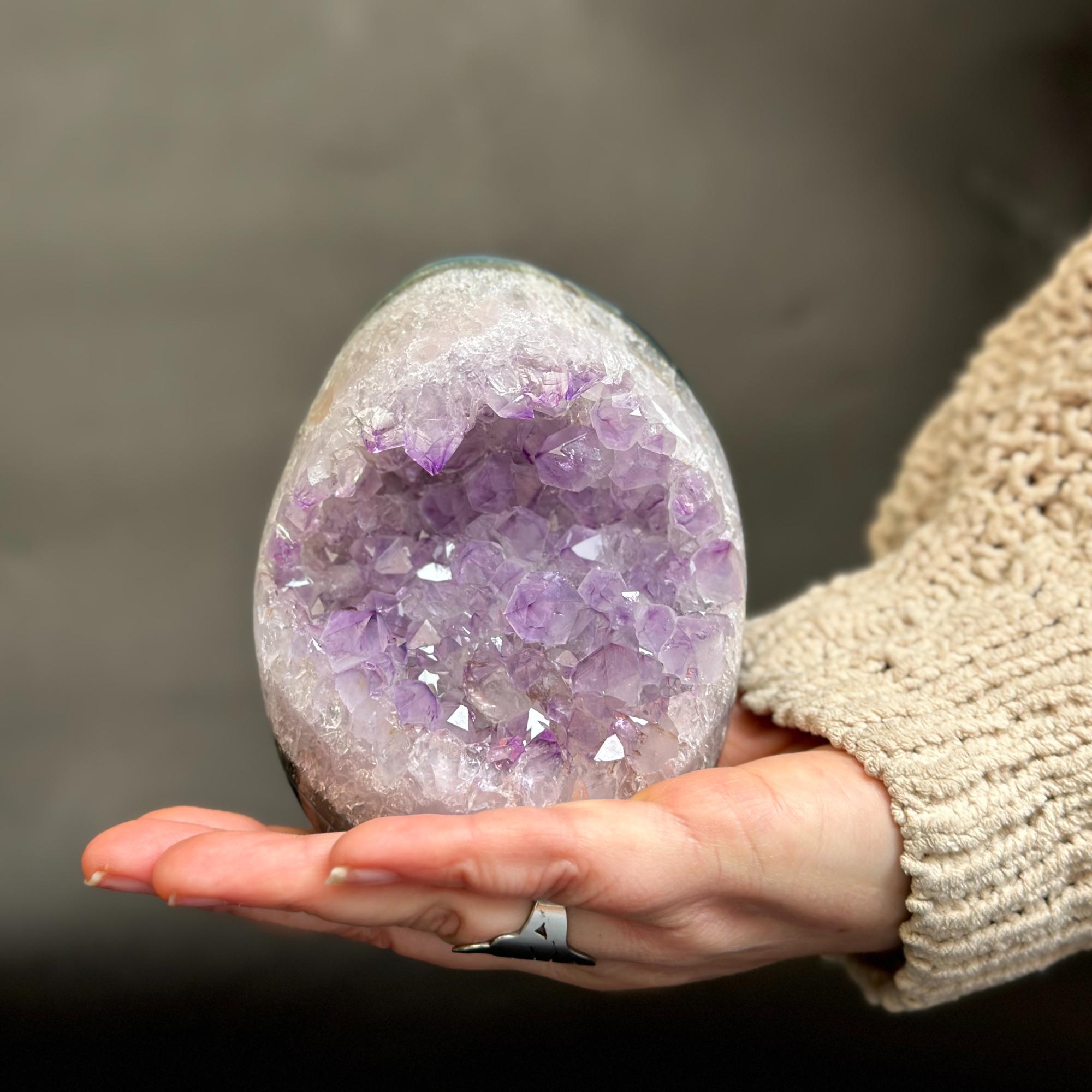 A person's hand holding a polished purple amethyst geode cluster, with a beige knit sweater sleeve visible against a dark gray background.
