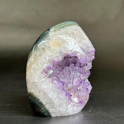 A polished natural crystal geode specimen with a prominent cluster of purple amethyst points on a translucent white and gray outer matrix, photographed on a dark surface against a neutral gray background.