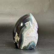 A polished agate stone sculpture with layered bands of dark black, white, lavender, blue, and metallic gold, displayed on a dark gray surface against a textured gray background.