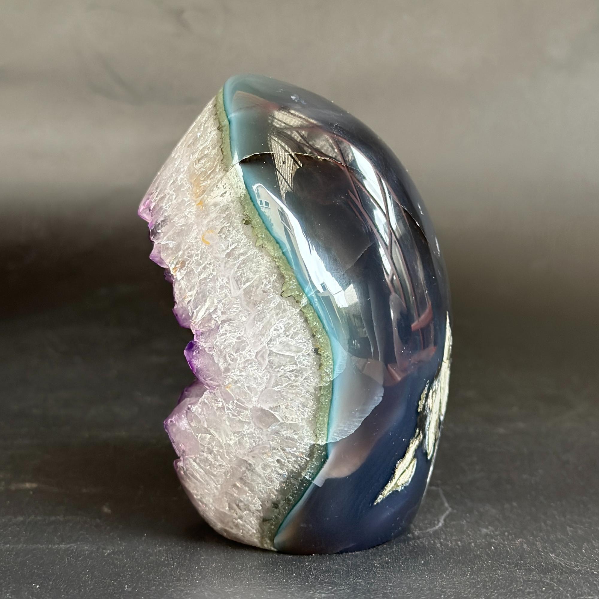 A polished geode specimen with a smooth, glossy dark blue and black outer shell, a light blue band, a white crystalline interior, and purple crystal formations along the left edge, set against a dark gray background.
