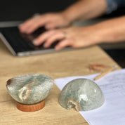 Two polished quartz half-spheres on a carob wood base, with one used as a paperweight, placed on a desk with a person typing on a laptop in the background.