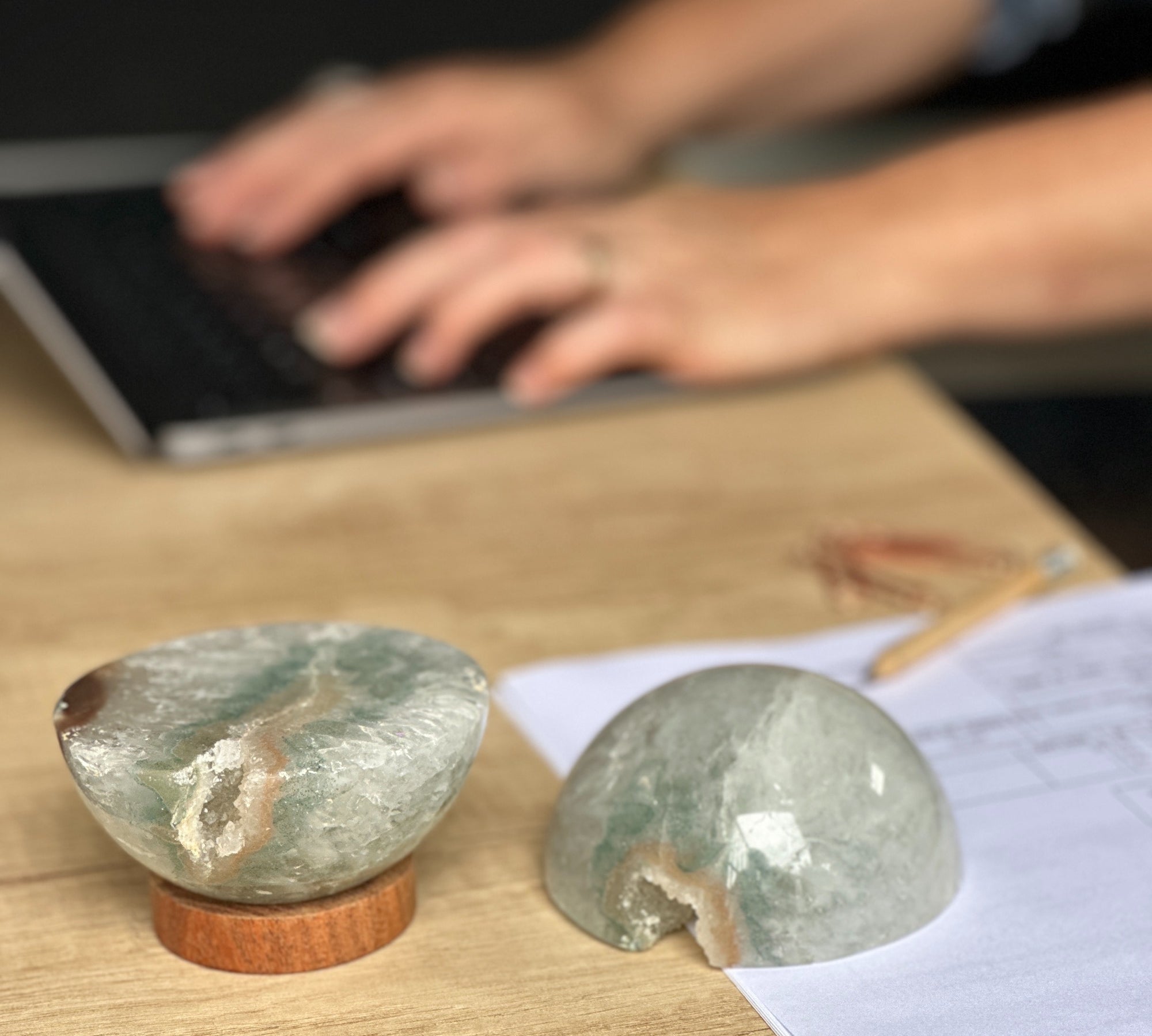 Two polished quartz half-spheres on a carob wood base, with one used as a paperweight, placed on a desk with a person typing on a laptop in the background.