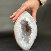 A hand holding a polished geode-shaped crystal sculpture with a white outer rind and a translucent lavender-purple crystal interior, set against a dark gray background.