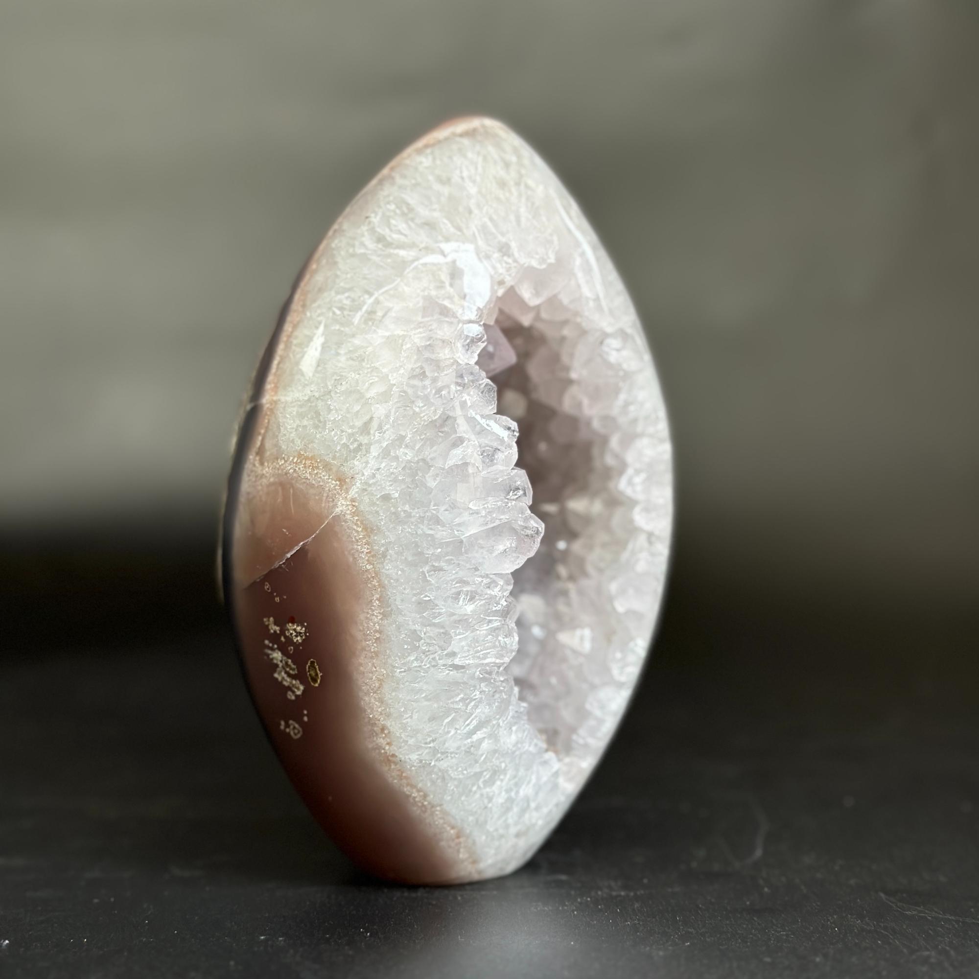 A polished geode-style decorative stone sculpture with a white crystalline interior and a smooth taupe-brown outer shell, standing upright on a dark surface against a soft gray background.