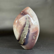 A polished agate stone with a teardrop shape, featuring a prominent metallic gold vein running vertically through a mottled mix of brown, purple, and beige tones, set against a dark gray background.