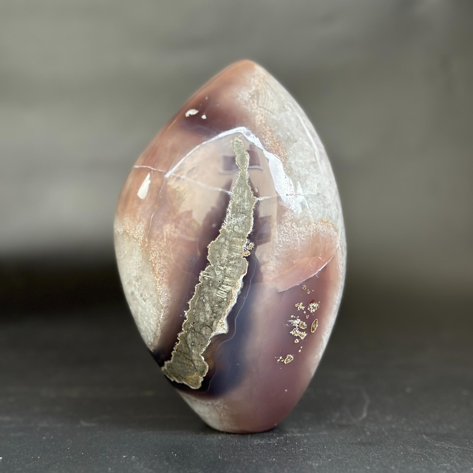 A polished agate stone with a teardrop shape, featuring a prominent metallic gold vein running vertically through a mottled mix of brown, purple, and beige tones, set against a dark gray background.