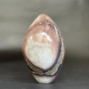 A polished agate stone sculpture with a teardrop shape, featuring a white crystal-like center and bands of brown, purple, and gray agate, standing on a dark surface against a gray background.