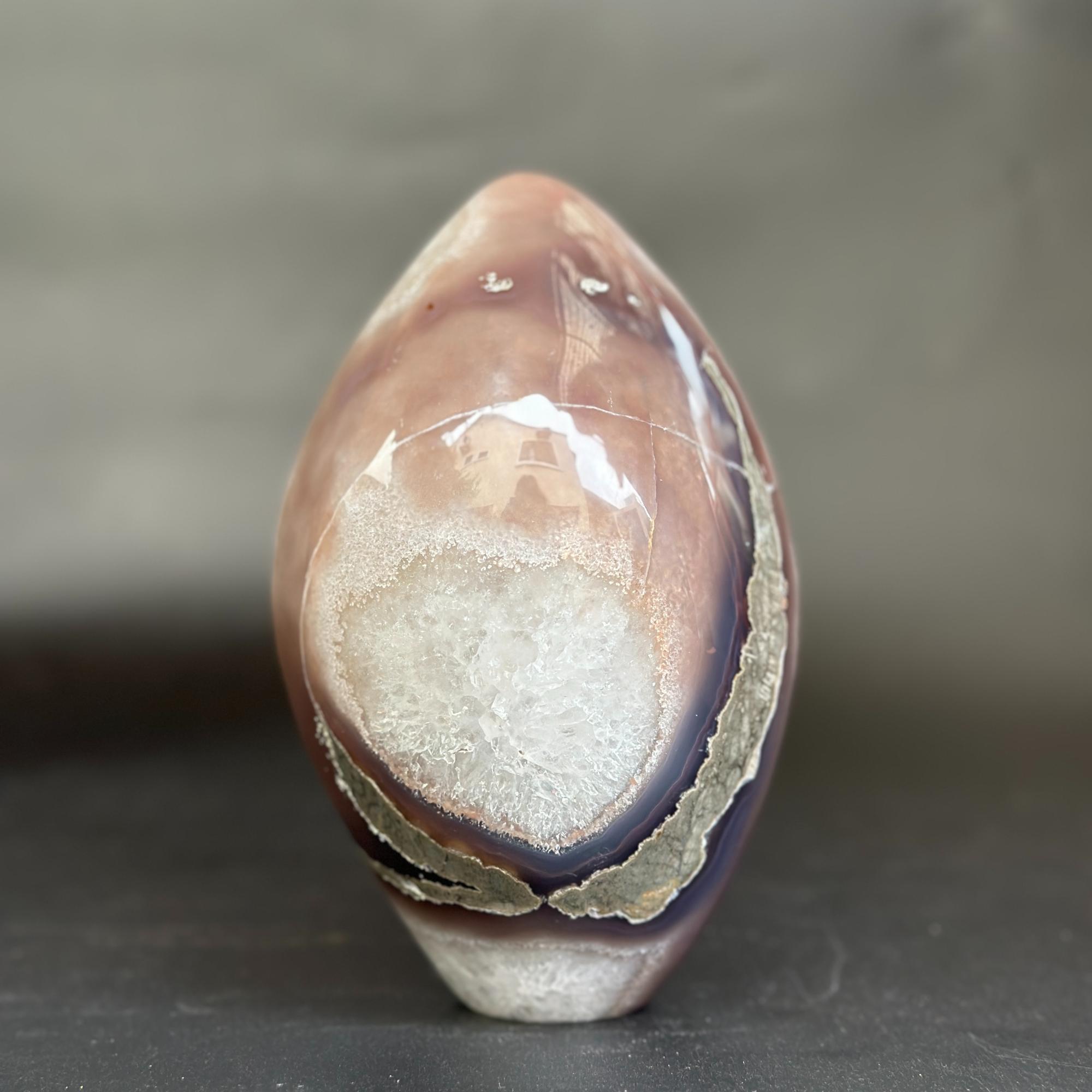 A polished agate stone sculpture with a teardrop shape, featuring a white crystal-like center and bands of brown, purple, and gray agate, standing on a dark surface against a gray background.