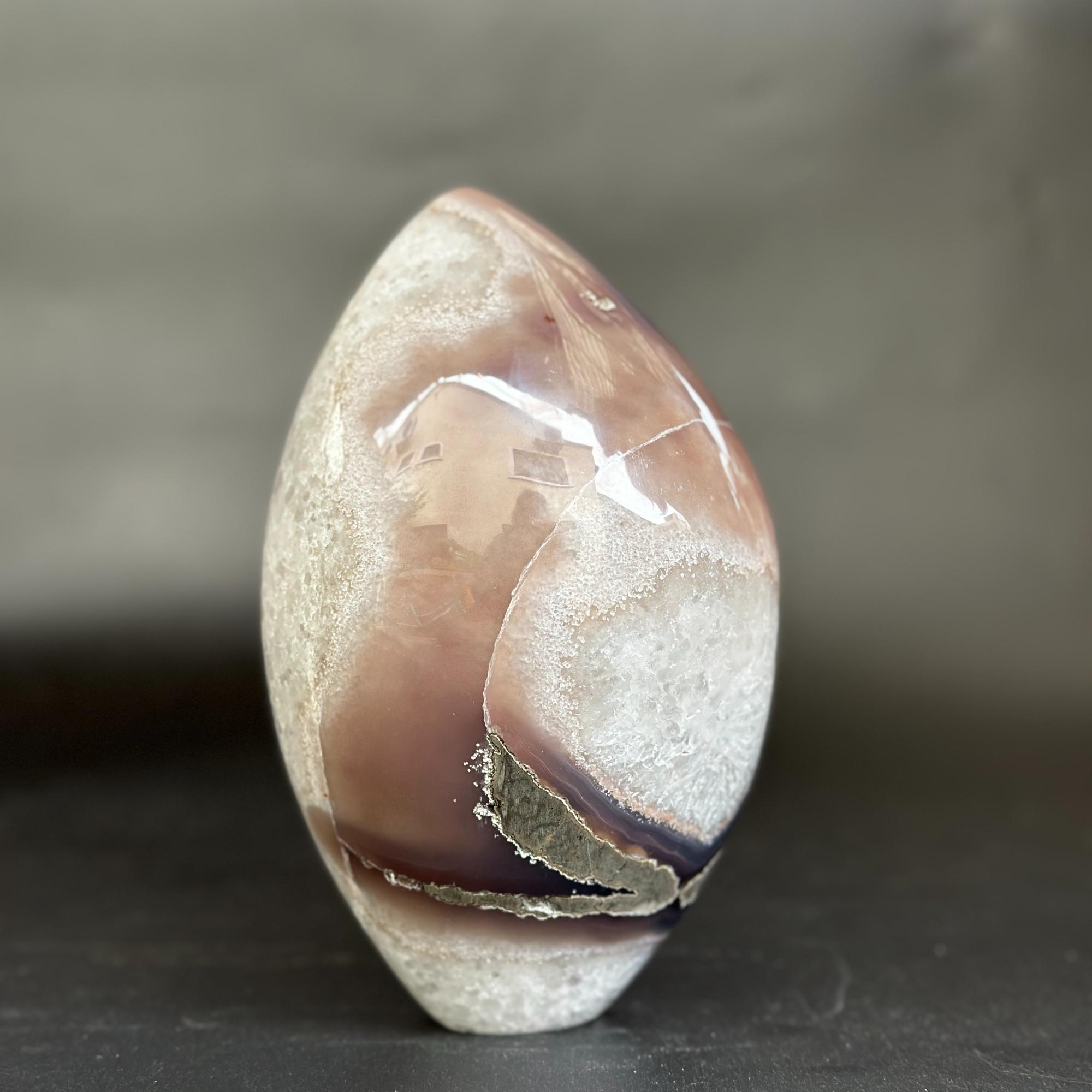 A polished agate crystal stone with concentric bands of brown, purple, and white, standing upright on a dark surface against a blurred gray background.