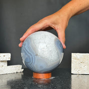 Hand touching a large, polished quartz and agate sphere with gray and white layers on a wood base.