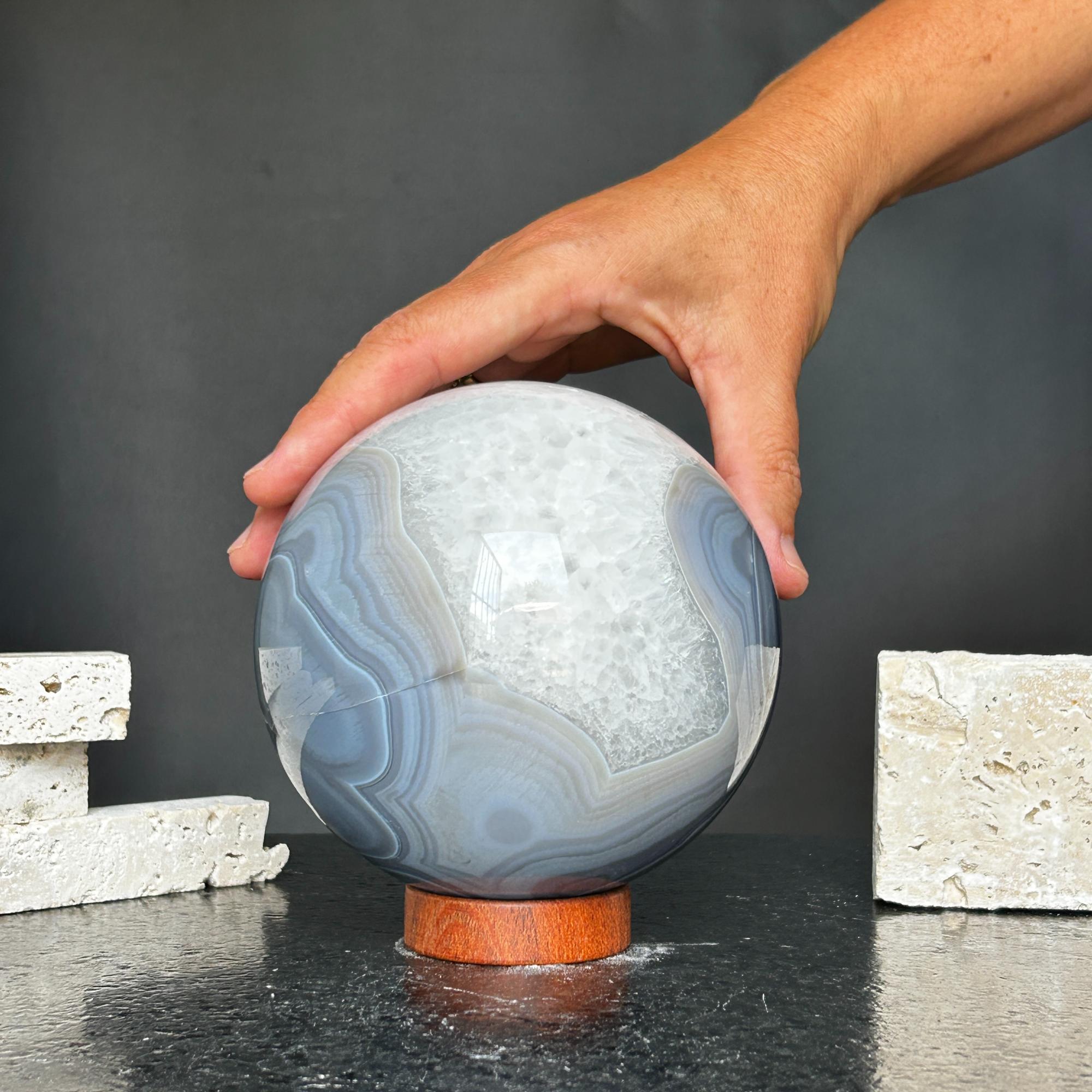 Hand touching a large, polished quartz and agate sphere with gray and white layers on a wood base.