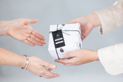 Hands exchanging a white gift box wrapped with black string and crystal patterns, featuring a 'FROM: GRACE' tag.