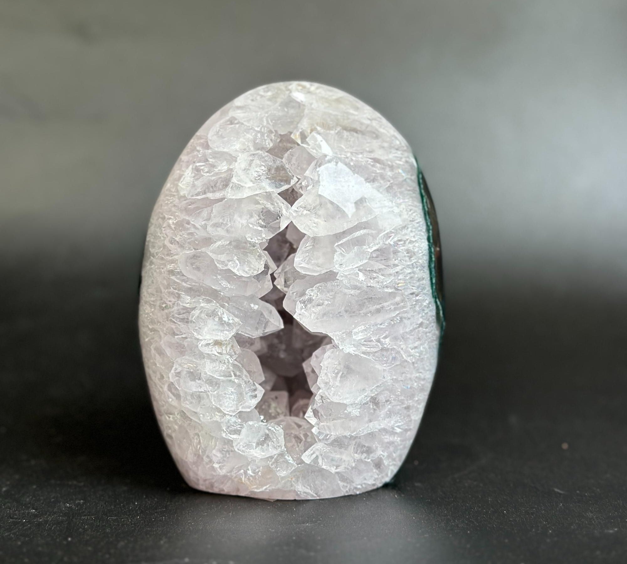 A polished geode-shaped crystal stone with a white and pale pink outer cavity and a dark interior, photographed on a dark gray surface against a gray background.